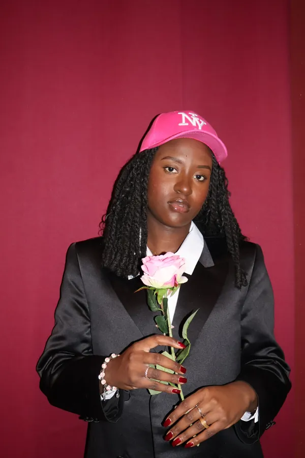 Portrait studio d'une membre de MTL Students sur fond cramoisi, casquette rose, veste noire et rose à la main.