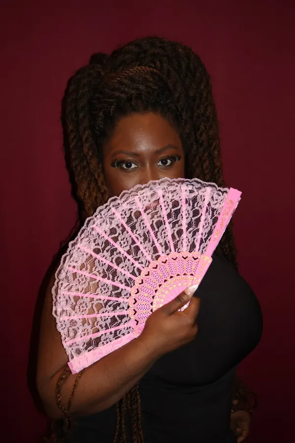 Portrait studio sur fond cramoisi : une membre de l'équipe cache son visage derrière un éventail en dentelle rose.