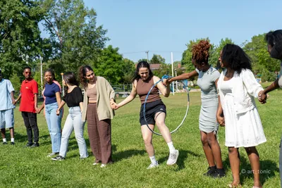 Une dizaine d'étudiants de MTL Students se tiennent par la main en cercle sur une pelouse ensoleillée et font passer un cerceau sans se lâcher.