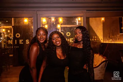 Trois étudiantes posent ensemble en soirée devant les baies vitrées éclairées d'un resto-pub.