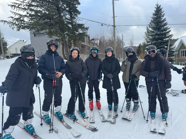 Un groupe d'étudiants de MTL Students en tenue de ski, casques sur la tête et bâtons en main, aligné dans la neige devant des sapins.