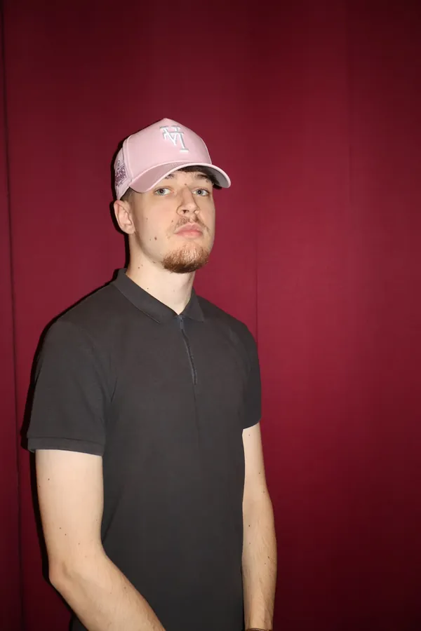 Portrait studio d'un membre de MTL Students en polo noir et casquette rose sur fond cramoisi.